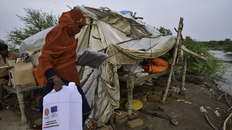 a woman in a red headscarf holds a water container in front of a makeshift shelter