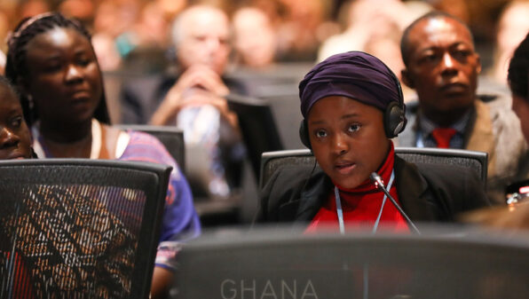 A girl in a purple headwrap speaks into a microphone at a desk with a Ghana badge