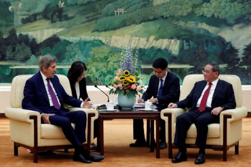 John Keery sitting in front ot Chinese premier Li Qiang in a hall.